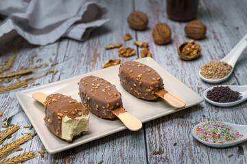 Bitten vanilla ice cream bar with chocolate and nut coating on ceramic plate. Rustic wooden background, surrounded by natural ingredients