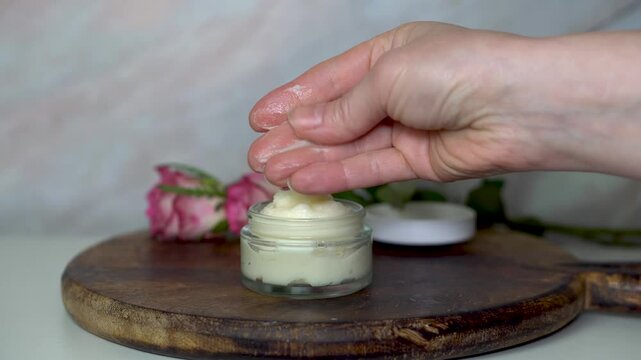 Woman testing the texture of homemade cream from a clear glass jar. Set on a dark wooden board with pink roses in background. Natural skincare, beauty routine, and wellness concept.