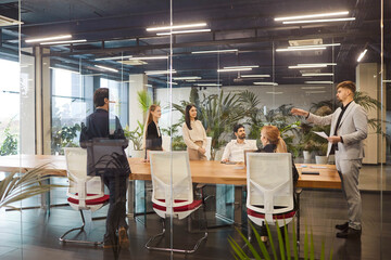 Diverse millennial business team briefing, talking in modern meeting room, negotiating on project behind transparent glass wall panoramic window, discussing deal in open space, modern office interior