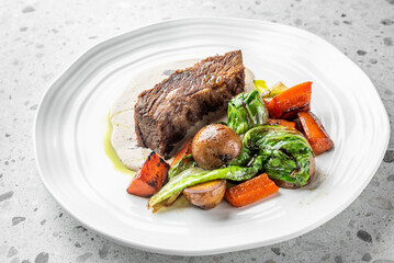 A succulent piece of braised beef served with roasted vegetables and a creamy sauce, elegantly plated on a white dish, perfect for showcasing gourmet dining and hearty meals.
