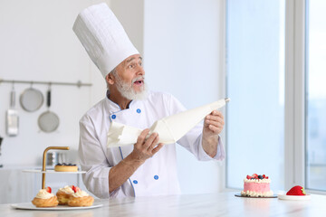 Funny confectioner holding pastry bag at table with desserts in kitchen