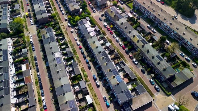 Aerial view of a residential neighborhood with rows of houses, solar panels on rooftops, parked cars along narrow streets, and small gardens, captured on a sunny day.