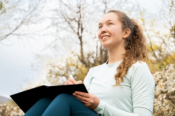 Obraz premium Dreaming young woman writing in notebook outdoors, relaxing, keeping diary, making wish list, sitting in nature park, taking notes, journaling, planning, drawing, fantasize, social detox, mindfulness