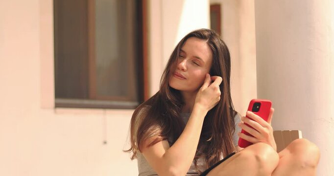 Young woman takes small wireless earbuds from charging case and puts headset in. Listen to music through new technology. Happy brunette woman sitting outdoor near house hold mobile phones.