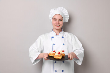 Confectioner in uniform holding delicious profiteroles on light grey background