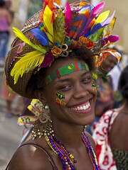 A brazilian woman adorned with a vibrant, colorful headdress featuring feathers and leaves, and wearing a variety of beads and necklaces. 