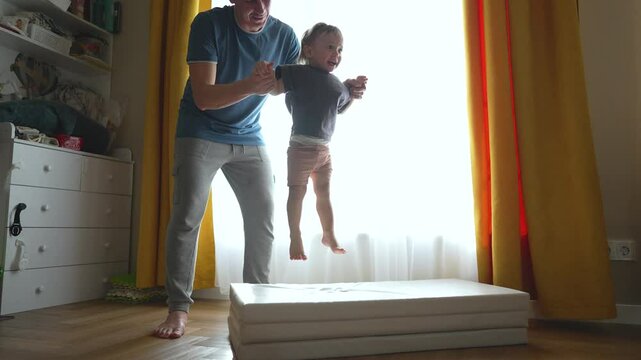 Father lifts son up jumping. Baby jumps on mattress with father. Happy baby enjoys jumping play. Dad and son bonding moment. Joyful baby jumps, father smiles. toddler jump fun on mattress with dad.