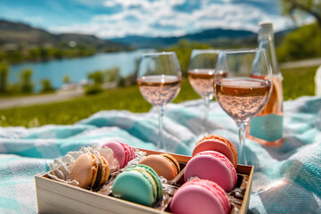 Close-up of colorful macarons in wooden box, beside rose wine glasses on blanket outdoors, scenic lake background, idyllic picnic scene