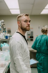 Fototapeta premium A scientist in a white lab coat holds a tablet, looking over his shoulder in a lab setting. Another scientist is blurred in the background.