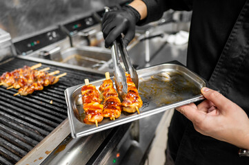 A chef in a black apron using tongs to place glazed chicken skewers onto a tray, showcasing the art of grilling in a bustling kitchen environment, perfect for highlighting culinary skills.