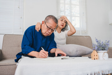 Elderly couple sitting on sofa with man focused on writing while woman looks worried and stressed...