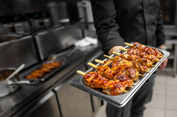 A chef in a black shirt holds a tray of glazed chicken skewers, freshly grilled and ready to serve, showcasing delicious barbecue artistry in a professional kitchen setting.
