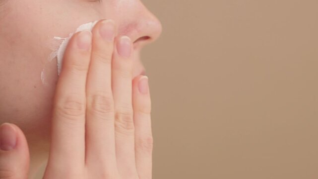 Close-up of a woman applies moisturizer, face cream or SPF sunscreen to cheek. Young girl applying foundation makeup in studio. Morning skincare routine. Concept ads for cosmetic products. Copy space