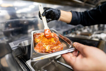 A chef in black gloves brushes marinade onto a piece of raw meat held in a stainless steel tray, highlighting the process of flavor infusion in a professional kitchen.