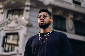 Confident young Black man in modern jewelry looking upward with focus, standing against elegant...