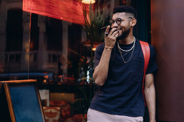 Confident young African American man stares into the camera with a slight smile. He wears a red backpack, dark t-shirt, pearl necklace and glasses on a city street at sunset