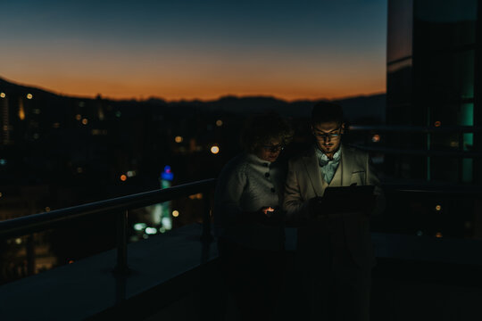 Two business colleagues collaborate on a project using a tablet during an evening brainstorming session on a rooftop. The city skyline and sunset provide a dramatic backdrop. - Powered by Adobe