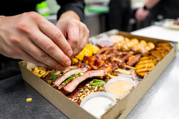 A chef's hands carefully garnish a platter of smoked meats with fresh herbs and sauces, showcasing a delicious assortment in a takeout-style box in a bustling kitchen.