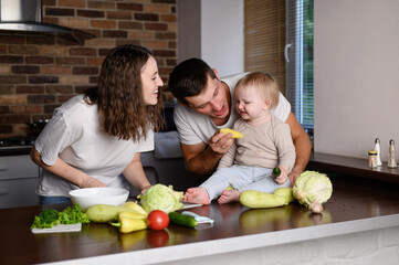 Family with baby busy cooking with fresh vegetables. Mom, dad busy cooking dinner, cutting vegetables, baby sitting on countertop and watching it. Concept of cooking healthy food at home