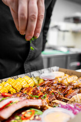  A chef sprinkles fresh herbs over a colorful platter of grilled meats, corn, and sides in a takeout box, enhancing the dish's flavor and presentation in a modern kitchen setting.