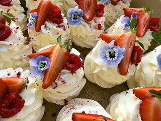 Mini pavlova desserts topped with whipped cream, strawberries, raspberries, and edible flowers,...
