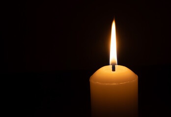 A single burning candle flame or light glowing on a big white candle isolated on black or dark background on table in church for Christmas, funeral or memorial service