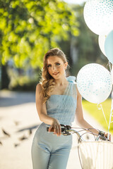 Young woman with balloons poses near bicycle in a bright outdoor park setting during daytime