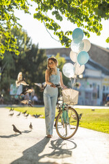 Young woman with balloons enjoys a sunny day in a park while standing by a bicycle surrounded by birds