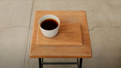 A simple cup of black coffee served on a wooden tray atop a rustic stool, captured in a minimalist setting with natural light and warm tones.