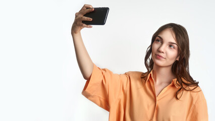 Young caucasian female taking selfie with smartphone in orange shirt against white background