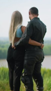 Man driver with beloved girlfriend embraces near modern motorcycle standing on hill top grass against calm lake on spring day backside view slow motion