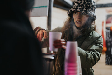 Woman pours a hot beverage into a cup, interacting with others during a winter event, emanating warmth and community. This setting highlights togetherness and generosity in a cozy outdoor environment.