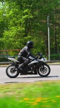Biker in black uniform and hardhat rides powerful motorcycle along large road past green park in city on sunny day side view slow motion