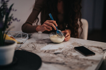 A woman sits at a marble table, savoring a dessert using a spoon. Her smart phone is nearby, creating a cozy and relaxed home atmosphere.