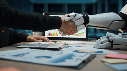 A human and robot engage in a handshake, representing collaboration in a modern office. This image...