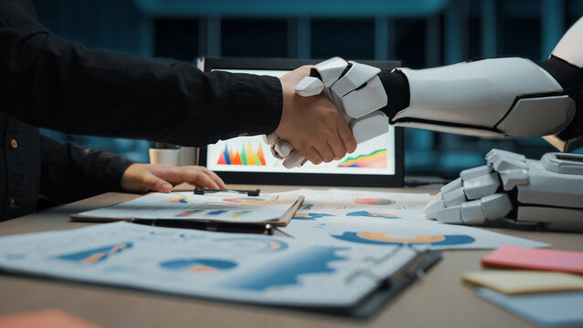 A human and robot engage in a handshake, representing collaboration in a modern office. This image captures the essence of technology working alongside people effectively.Noogenesis