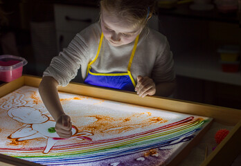 Beautiful little girl draws with sand on a light square, art therapy, sand animation, creative leisure for children, selective focus	
