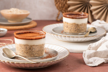 Creamy tiramisu overnight oats and chia pudding topped with yogurt and cocoa powder, served in elegant glass cups on white plates with spoons, set on a rustic table