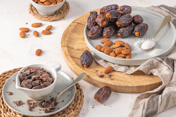 Medjool dates, almonds and spoonful of shredded coconut on a plate with a wooden board, surrounded by scattered almonds, dates, and chocolate chunks, creating a rustic and appetizing scene