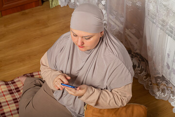 Muslim woman in gray hijab in room on plu on pillows scrolls corresponds