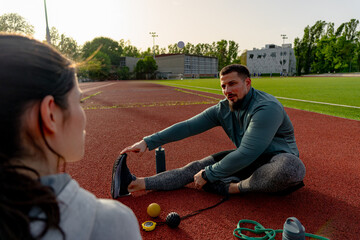 Male athlete stretching legs on running track with personal trainer assisting and giving advice on running technique