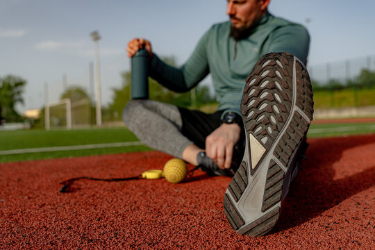 Male athlete taking a break after training on a running track, holding a water bottle and stretching his legs - Powered by Adobe