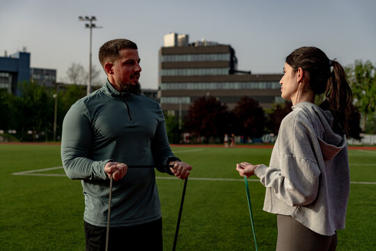Personal trainer assisting a woman performing exercises with resistance bands on a sports field, promoting fitness and active lifestyles