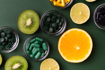 Many dietary supplements, vitamins and minerals, colored pills and tablets in small bowls, kiwi, lime and orange as a source of natural vitamins from above on green background. Vitamins and fruits. 