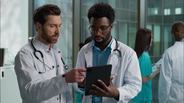 Multiethnic doctors conversation African American therapist and Caucasian surgeon physician men talking brainstorming diagnosis holding tablet device thinking difficult case at hospital clinic hallway