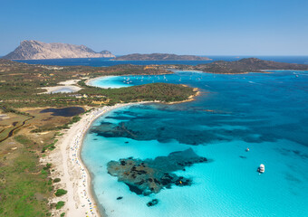 Aerial view of white sandy beach with umbrellas, luxury yachts and boats on blue sea, beautiful island on summer sunny day. Sea coast, turquoise transparent water in Sardinia, Italy. Top drone view