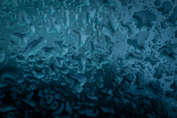 Close-up view of water droplets forming abstract patterns on a blue-tinted glass surface, creating a moody, textured background with copy space.