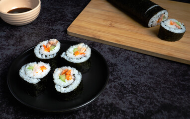 A plate of sushi sits on a table next to a cutting board. The sushi is cut into pieces and arranged on the black plate. Black background