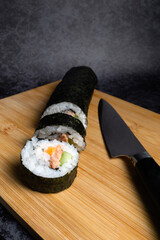 A sushi roll with a knife on a wooden cutting board. The knife is on the right side of the sushi roll. Black background
