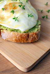 a slice of toasted bread topped with mashed avocado, a fried egg and fresh green cress springs on a wooden board